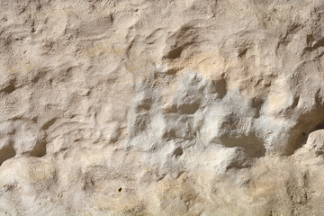 A close-up horizontal shot of an extremely rough and uneven wall texture with thick, weathered light beige stucco under harsh sunlight