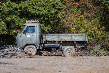 Obraz premium A weathered military-style flatbed truck rests on dry earth beside stacked firewood, framed by autumn forest foliage