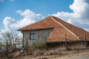 A weathered house with a red-tiled roof stands behind a wire fence in a dry, overgrown field beneath a partly cloudy autumn sky.