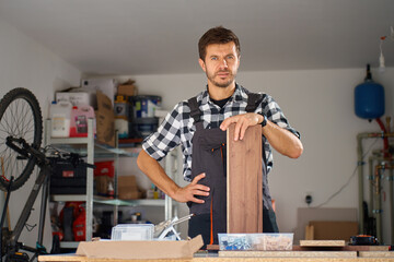Carpenter standing in home workshop with wooden panel and tools on workbench. Portrait of small business owner in garage. Concept of woodworking and custom furniture making