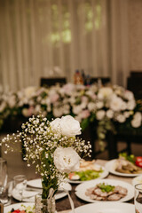 Fresh white roses standing in a small vase on a banquet table for decoration, among various dishes in a restaurant during a wedding celebration
