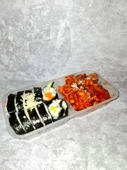 A plastic take-out container holding a Korean combo meal with sliced kimbap rolls topped with mayonnaise and grated cheese, alongside a portion of spicy red-sauced chicken (Dakgalbi or Yangnyeom).