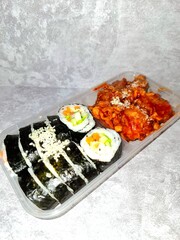 A plastic take-out container holding a Korean combo meal with sliced kimbap rolls topped with mayonnaise and grated cheese, alongside a portion of spicy red-sauced chicken (Dakgalbi or Yangnyeom).