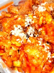 A close-up, mouthwatering shot of crispy Korean fried chicken (Yangnyeom or Gangjeong) pieces coated in a thick, glossy, sweet, and spicy red-orange glaze, heavily topped with white sesame seeds.