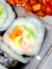 Blurred. A close-up view of sliced Korean kimbap or sushi rolls, showcasing the vibrant filling of rice, carrot, and cucumber wrapped in dark seaweed, with a pile of sesame seeds visible on the left.
