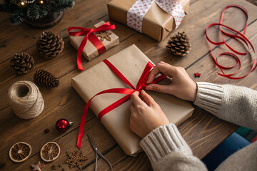 Wrapping a Christmas gift with red ribbon on a wooden table with decorations.