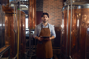 Confident man in an apron is holding a digital tablet inside a brewery