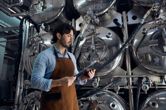 A man wearing an apron is holding a tablet inside a brewery facility