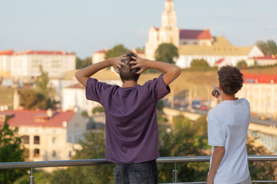 Two young males, one with hands on head and the other pointing, are observing a scenic city view from a balcony, capturing a moment of contemplation and discovery