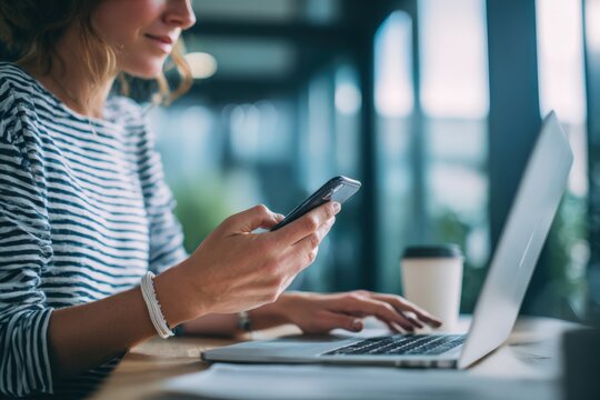 A woman is sitting at her desk using a laptop and cell phone