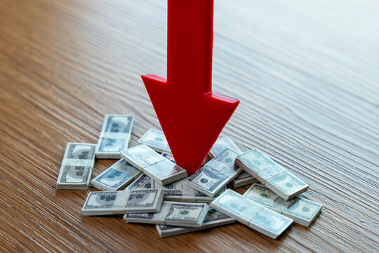 Red downward arrow pointing at piles of US dollar bills on wooden surface, representing financial loss, dollar depreciation, and economic downturn concept.