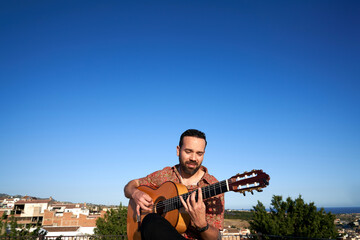 Flamenco guitarist playing acoustic guitar outdoors, concentrating on music in a Spanish village landscape
