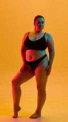 Standing plus size woman in black lingerie posing with neon light contrast on orange background. Concept of underwear branding, lingerie ads, empowerment campaigns, stylish editorial portraits.