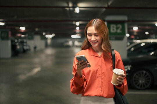 Smiling woman standing in parking garage using smartphone while holding coffee cup