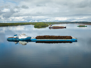 Large barge transporting logs through calm waters in forested area under cloudy sky