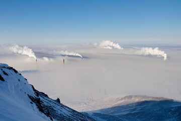 Steam rises from industrial smokestacks against a clear blue sky in a snowy mountainous landscape