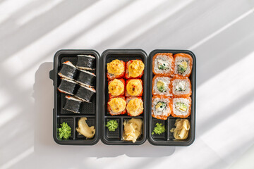 Assorted sushi rolls in black plastic containers with wasabi and pickled ginger on white background. Japanese food delivery or takeout concept, top view in natural light