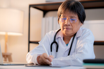 Senior asian doctor writing prescription or document on tablet in clinic office, managing patient record, healthcare and diagnosis, elderly female physician writing on tablet, medical concept.