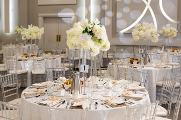 Served tables with white flowers in the center in the large hall create an elegant and festive atmosphere. Round tables with white tablecloths and cutlery are prepared for guests