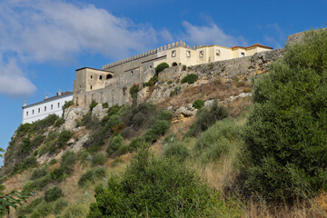 Fototapeta premium The rugged stone walls and historic buildings of Palmela Castle perched atop a steep, vegetation-covered hill in the Portuguese countryside.