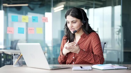Confident young businesswoman in a wireless headset talking on a video call using a laptop sitting at workplace in office. Female manager has business meeting, speaks remotely at an online conference - Powered by Adobe