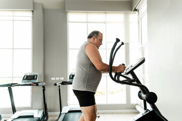 Senior man trains on elliptical in bright gym with large windows, sweating through cardio workout. Scene shows strength, motivation, discipline, rehabilitation, recovery, effort, and overall wellness.