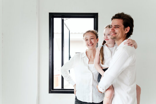 Fototapeta Happy family standing by a window in a bright, unfurnished room  father holding child and mother smiling, suggesting new home, renovation, or real estate success.