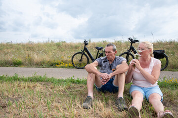 Senior couple enjoying a break from their bicycle tour, sitting relaxed on a grassy bank next to their e bikes, appreciating the outdoors and the simple pleasure of leisure and togetherness