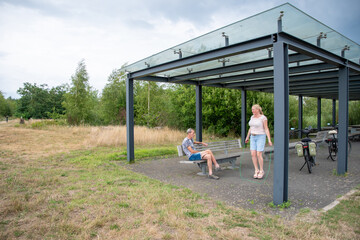 Mature woman skipping rope near touring bicycles while her partner relaxes on a park bench, enjoying active retirement, outdoor travel and healthy, energetic summer leisure