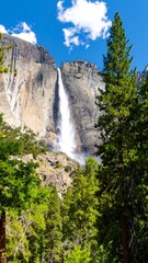 Waterfall cascading down granite cliff face, surrounded by lush green trees and a partly cloudy sky