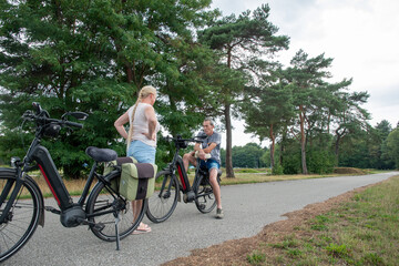 Fototapeta premium Senior man and woman stopping their electric bikes on a paved path, relaxing and conversing during a summer cycling trip, embracing a healthy and active lifestyle