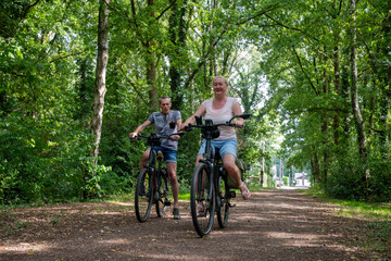 Naklejka premium Mature couple riding electric bikes on a dirt path, surrounded by lush green trees, enjoying a leisure activity and healthy lifestyle during their travel