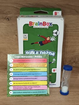 BrainBox soccer quiz game with colorful box, trivia card featuring questions about Legia Warsaw, and a sand timer on a wooden surface