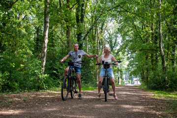 Mature couple standing with their bikes on a dirt path in a green forest, holding hands and sharing a moment of romance during a summer outdoor activity