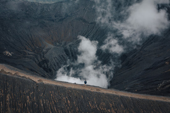 Aerial view of a lone figure standing before the Bromo volcano's caldera, a stark contrast of warm earth tones against the cool, smoky depths, Mt. Bromo, East Java, Indonesia.