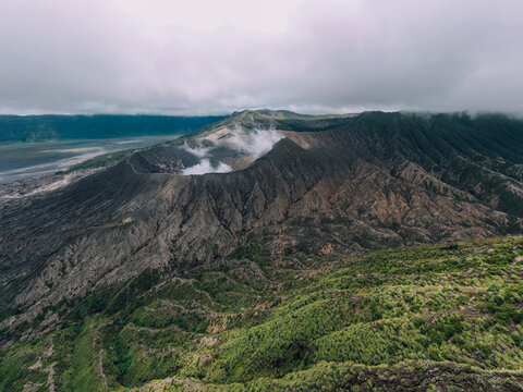 Aerial view of the rugged, smoky crater of Mt. Bromo, where dark volcanic rock meets vibrant green vegetation under a sky heavy with clouds, Mt. Bromo, East Java, Indonesia.