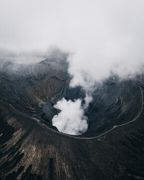 Aerial view of the stark, textured landscape and volcanic crater shrouded in ethereal white smoke, a mesmerizing dance of dark earth and light mist, Mt. Bromo, East Java, Indonesia.