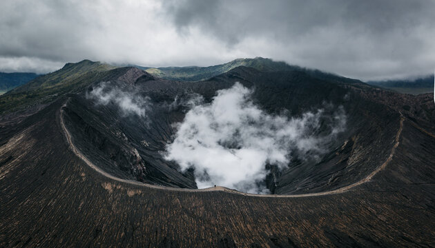 Aerial view of the Bromo volcano crater, a dark, textured landscape pierced by plumes of white smoke under a heavy, clouded sky, Mt. Bromo, East Java, Indonesia.