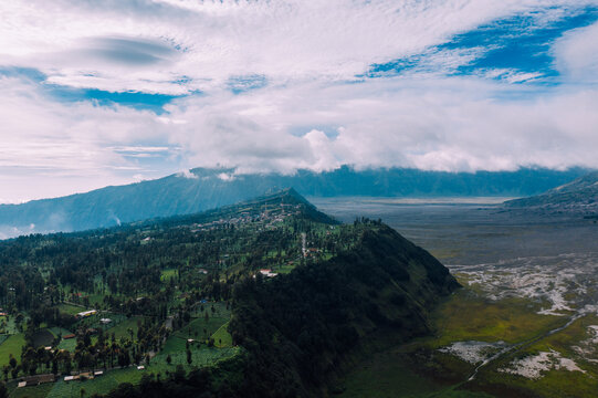 Aerial view of the vibrant green hills contrasting against the volcanic ash plains under a cloudy sky, Mt. Bromo, East Java, Indonesia.