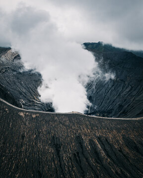 Aerial view of a volcanic landscape where the dark slopes meet a pale, smoky crater under a brooding sky, Mt. Bromo, East Java, Indonesia.