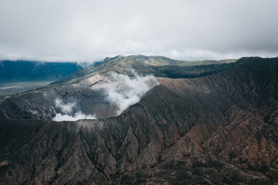 Aerial view of the rugged terrain of Mt. Bromo's volcanic caldera, where plumes of smoke rise against a backdrop of rolling green hills, Mt. Bromo, East Java, Indonesia.