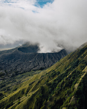 Aerial view of the majestic volcanic landscape, shrouded in ethereal clouds, where the rugged, dark crater contrasts with the vibrant green slopes, Mt. Bromo, East Java, Indonesia.