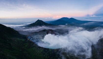 Aerial view of a volcanic landscape where the crater lake steams amidst the rugged terrain and distant peaks under a pastel sky, Mt. Ijen, Jawa Timur, Indonesia.