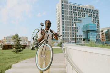 Black young adult man carrying bicycle up stairs outdoors near modern office building, wearing...