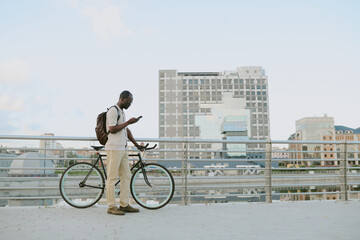 Young adult Black man standing outdoors next to bicycle using smartphone, wearing backpack, urban cityscape in background, appearing focused while interacting with device