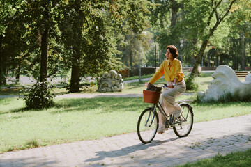 Middle aged Caucasian woman riding bicycle along park path, hands on handlebars, looking ahead, surrounded by green trees and grass, enjoying outdoor activity in urban park setting