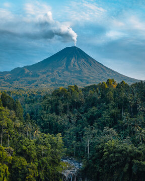 Aerial view of the majestic Semeru volcano rising above lush green jungle, with a trail of smoke ascending into the blue sky, Tumpak Sewu, Jawa Timur, Indonesia.