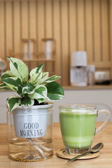 A cozy close-up of a green matcha latte in a glass cup beside a small indoor plant in a pot, placed on a light wooden table with a modern minimal kitchen background. Perfect for concepts of relaxation