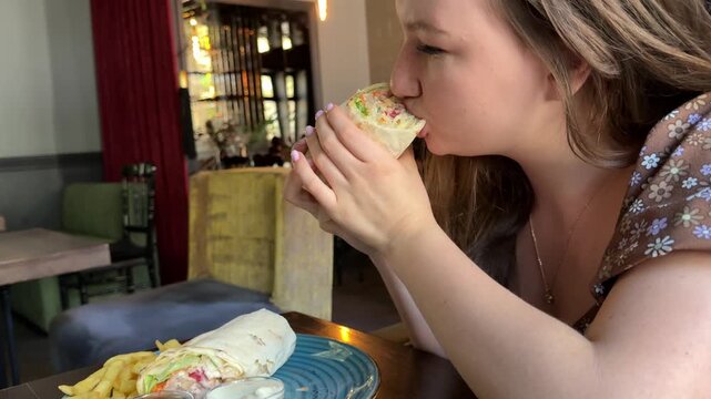Young blond attractive woman eating delicious wrap (burrito) in restaurant. High quality 4k footage