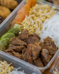 close-up of spiced braised beef chunks with vermicelli, bean sprouts and lime slices in a meal box, suitable for editorial food, menus, packaging and meal-prep visuals
